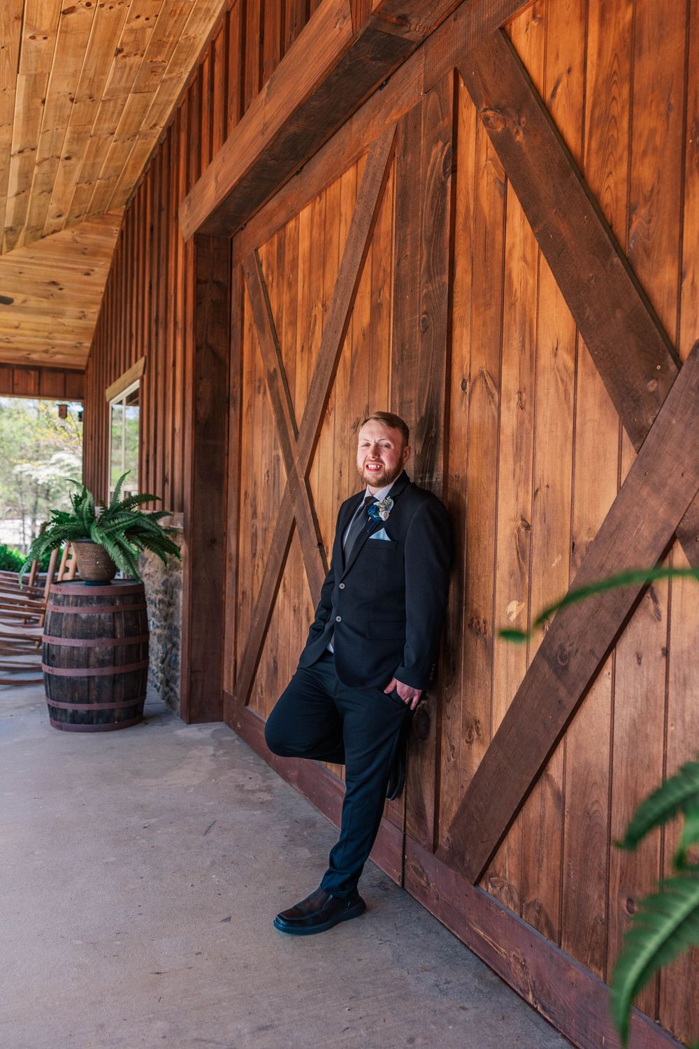 Groom waits in rustic wooden barn setting with vintage barrels and exposed beams.