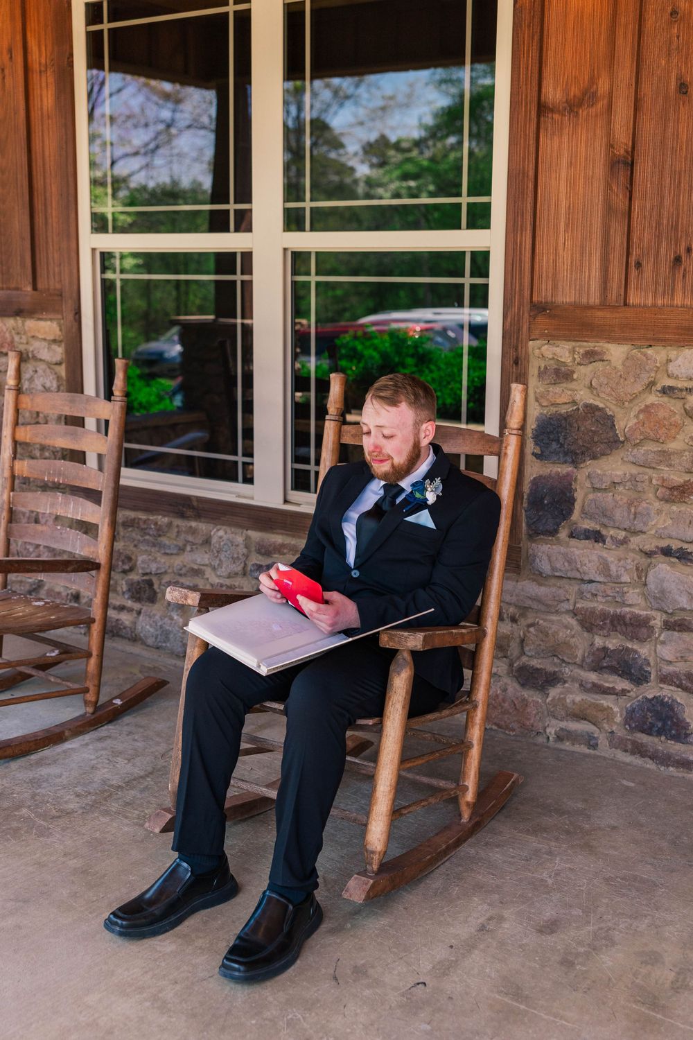 A person in formal attire sitting in a rocking chair on a stone porch reading.