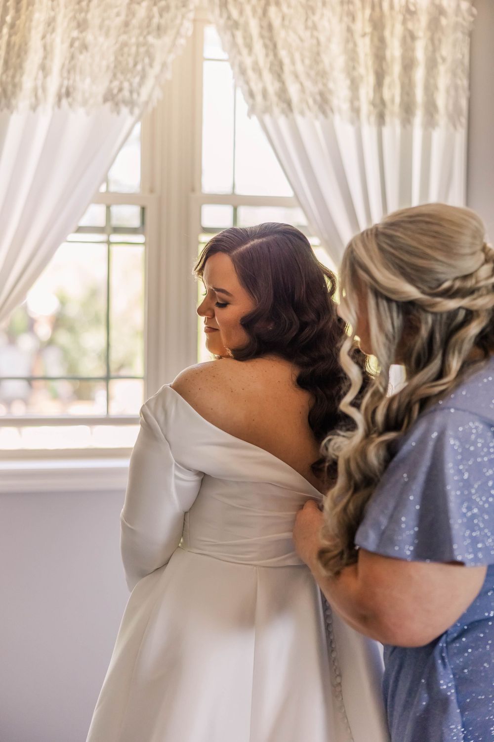 Wedding dress being zipped up in bright white room with flowing curtains.