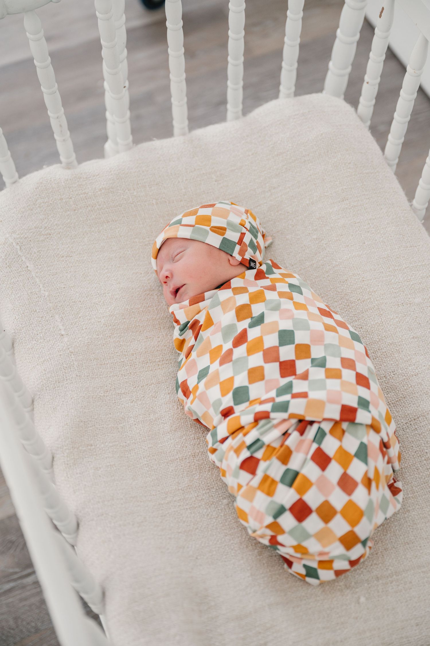 Baby swaddled in colorful checkered blanket sleeps peacefully in white wooden crib on cream bedding.