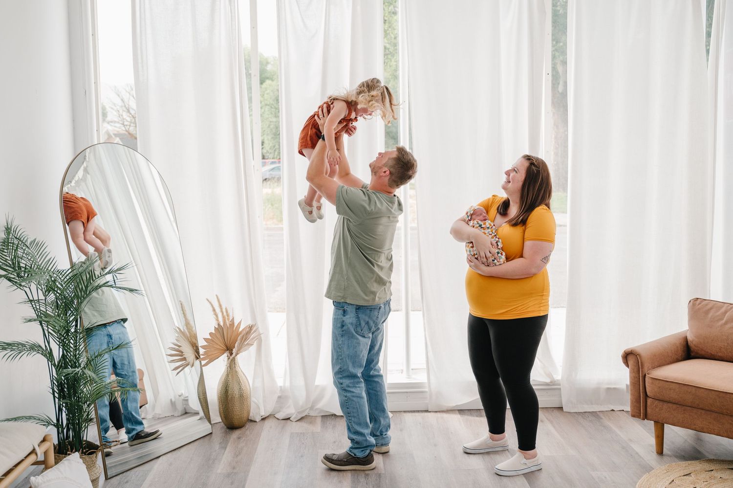 A family plays together in a bright living room with white curtains and potted plants beside a brown leather chair.