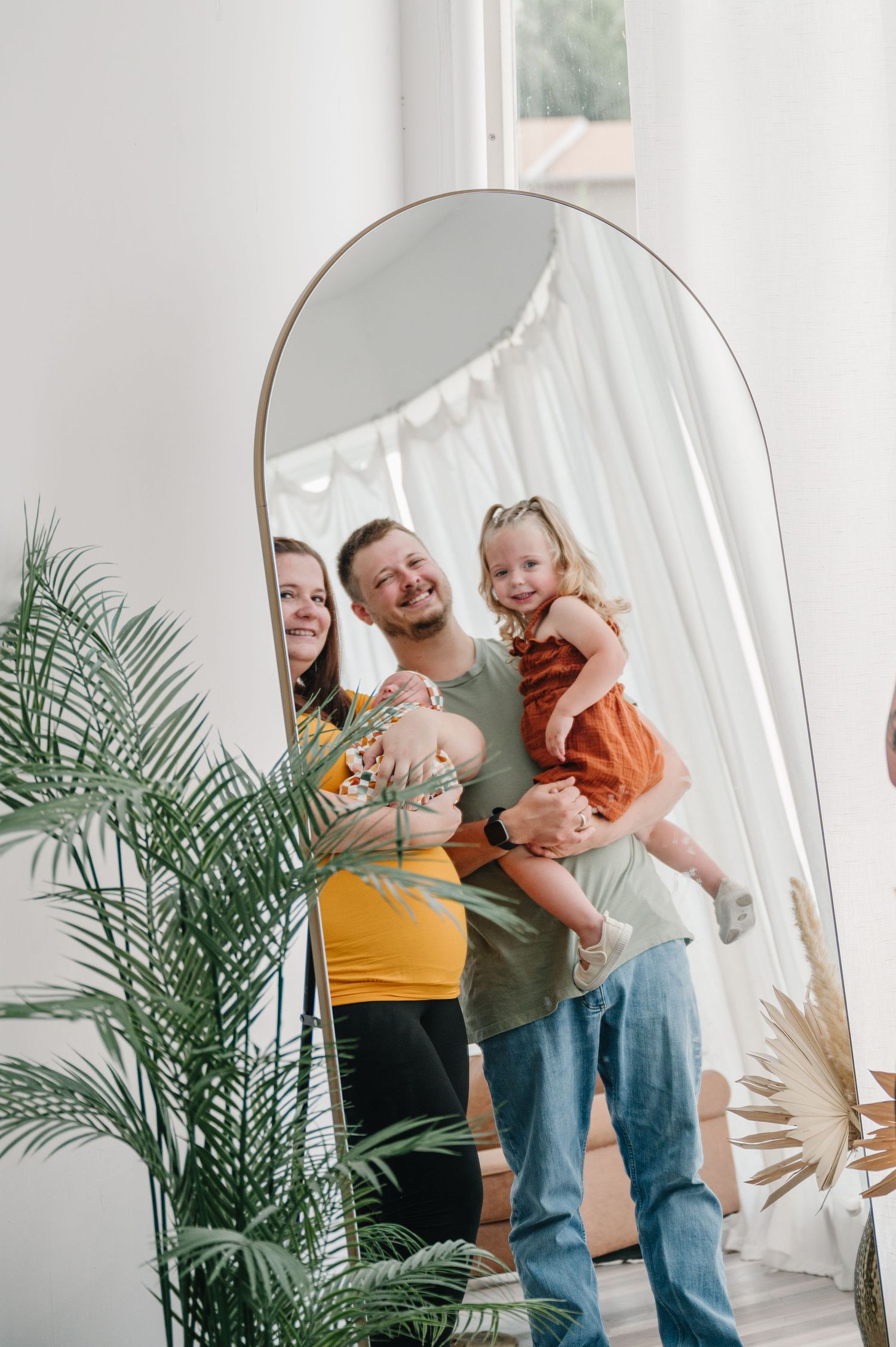A group reflects together in an arched mirror with a palm plant and white curtain in a bright minimalist setting.