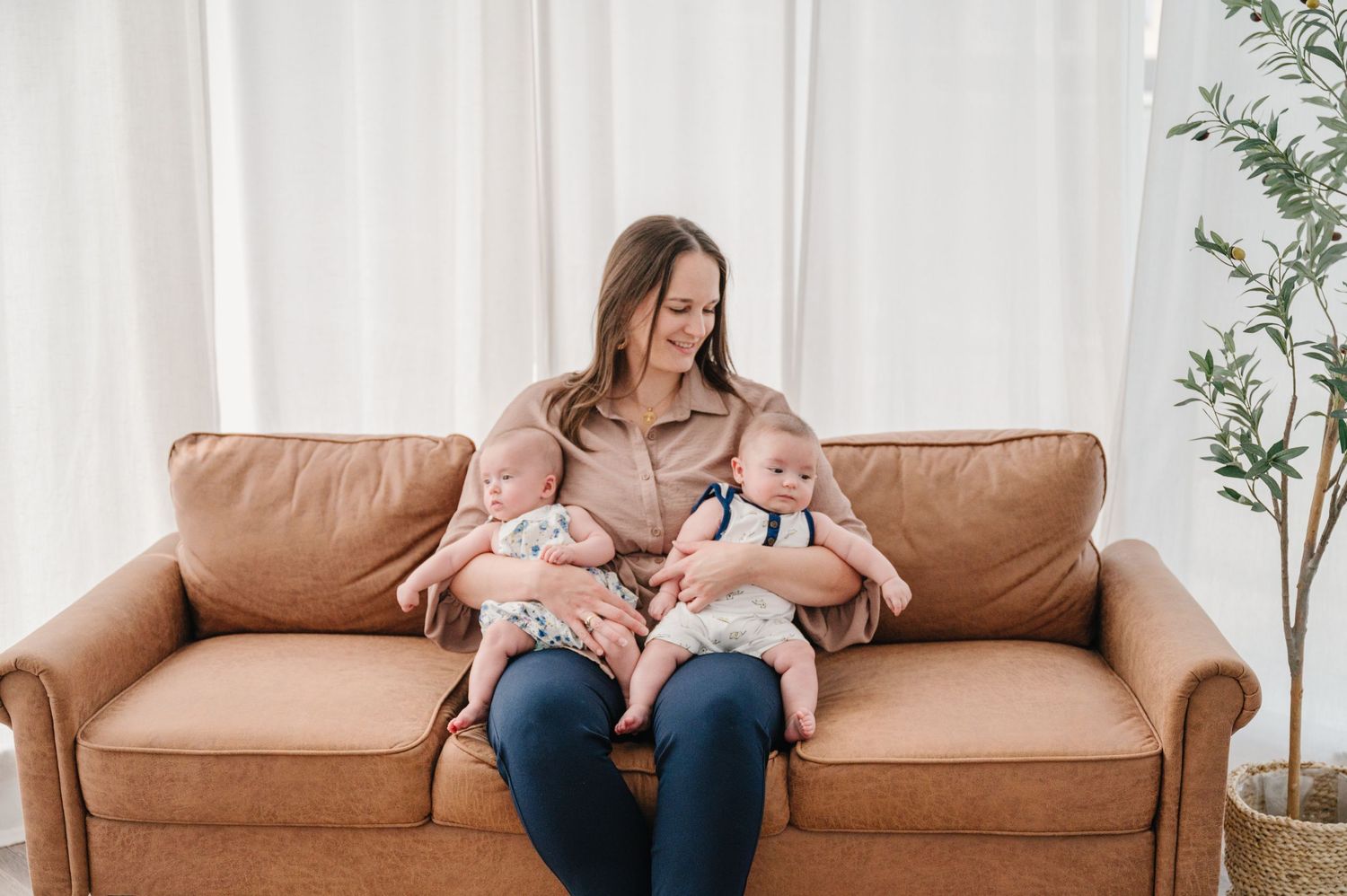 Woman sitting on tan leather couch breastfeeding twins in a bright, modern living room.