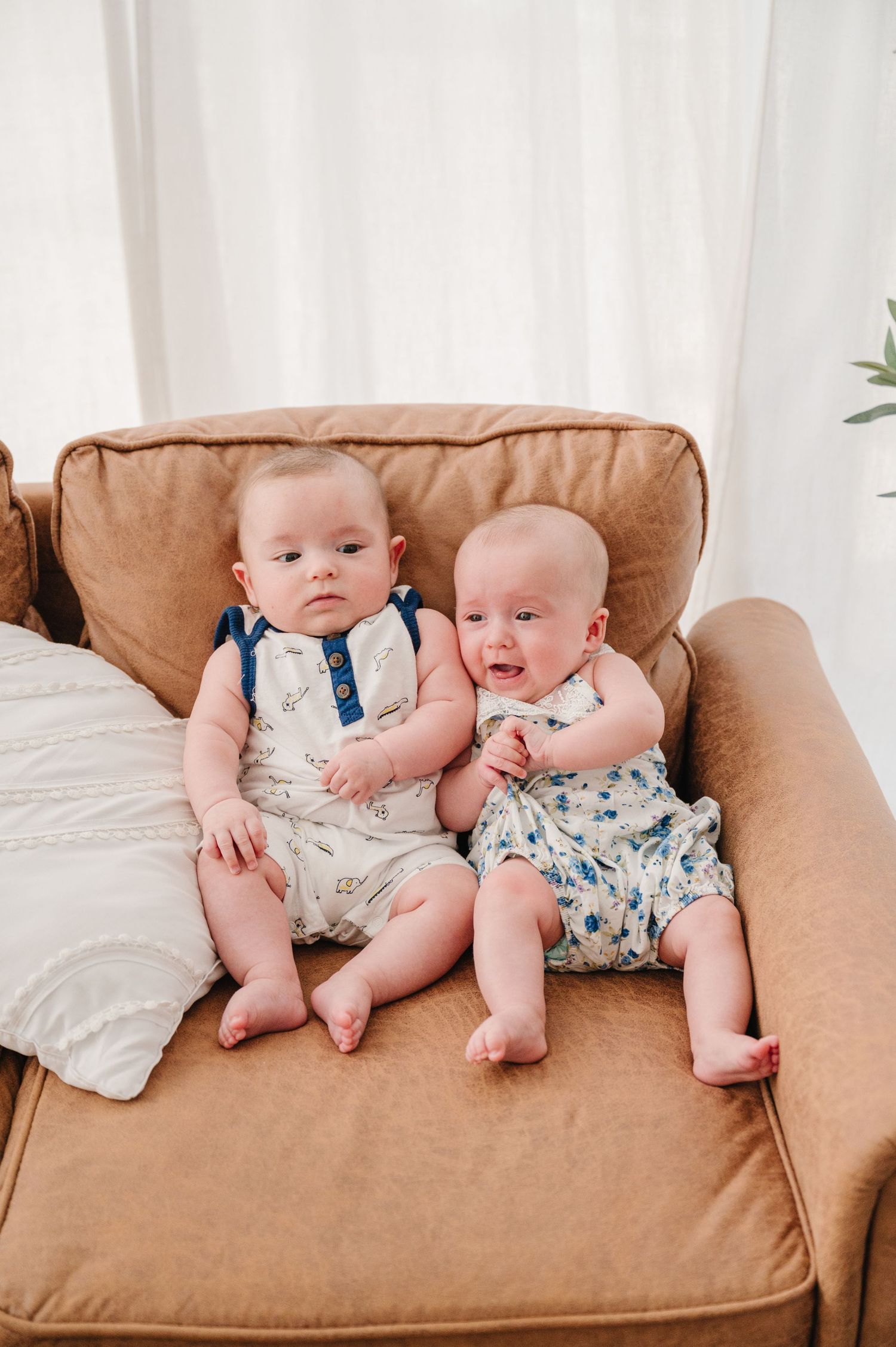 Two babies in coordinated floral outfits sit together on a tan leather couch in a bright room.