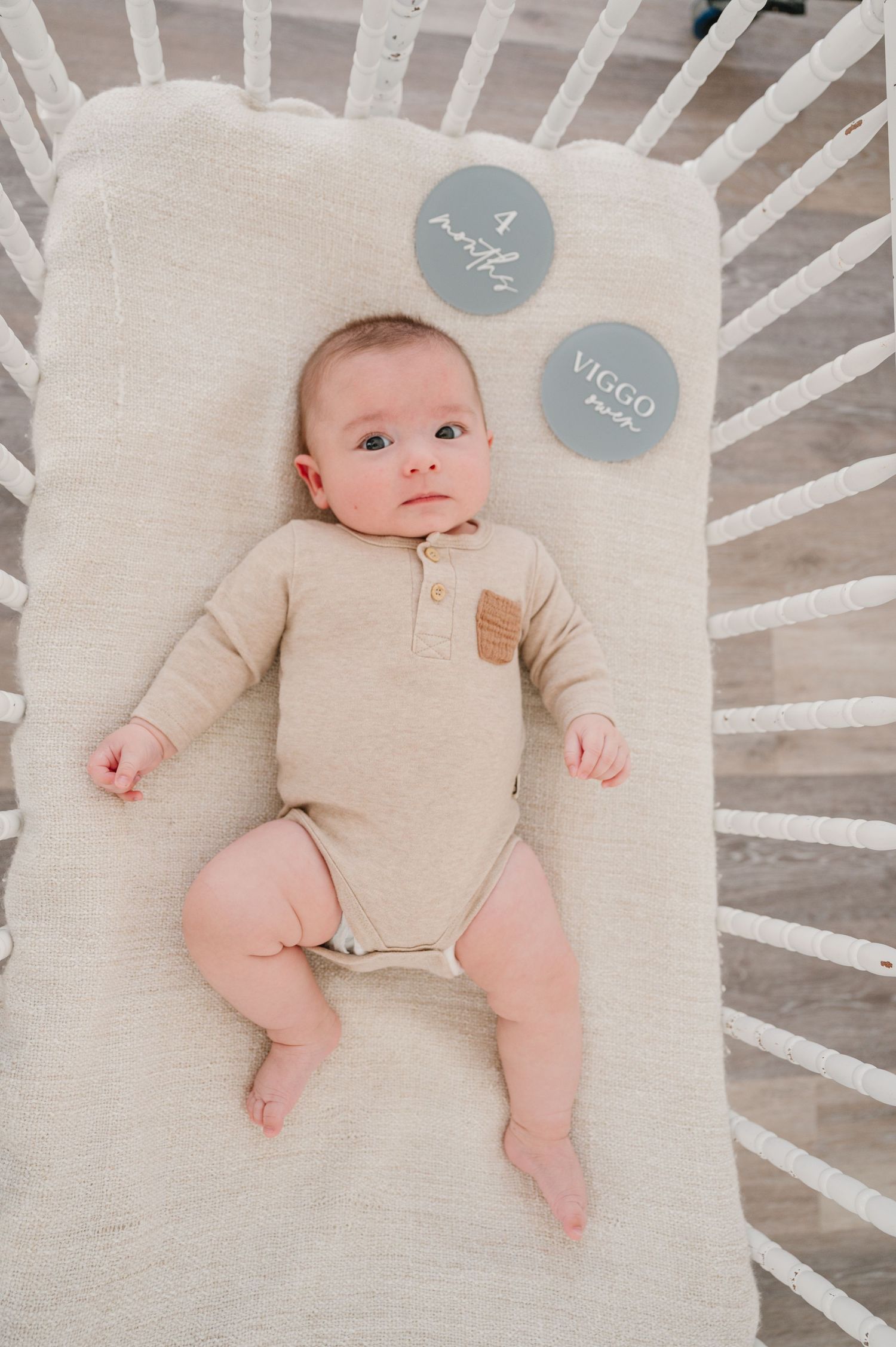 Baby wearing beige henley onesie lying on cream-colored crib mattress with two gray milestone markers.