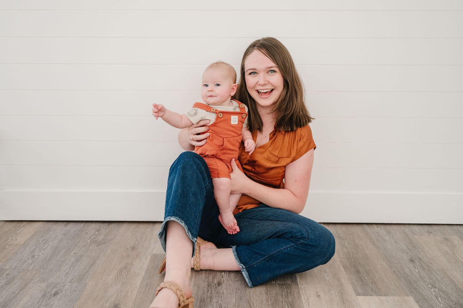 A pair in matching rust orange clothing sit and laugh together on a grey hardwood floor.