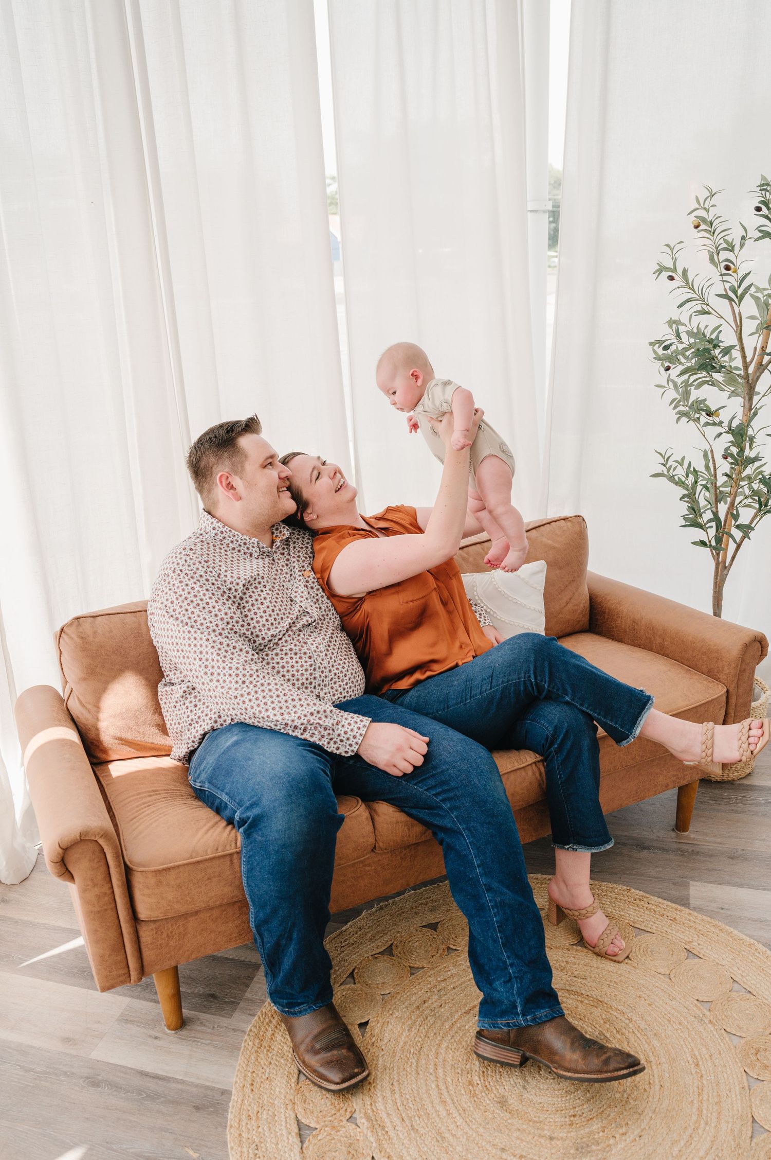 Family enjoying joyful moment together on a tan leather couch in a bright room with white curtains and potted plant.