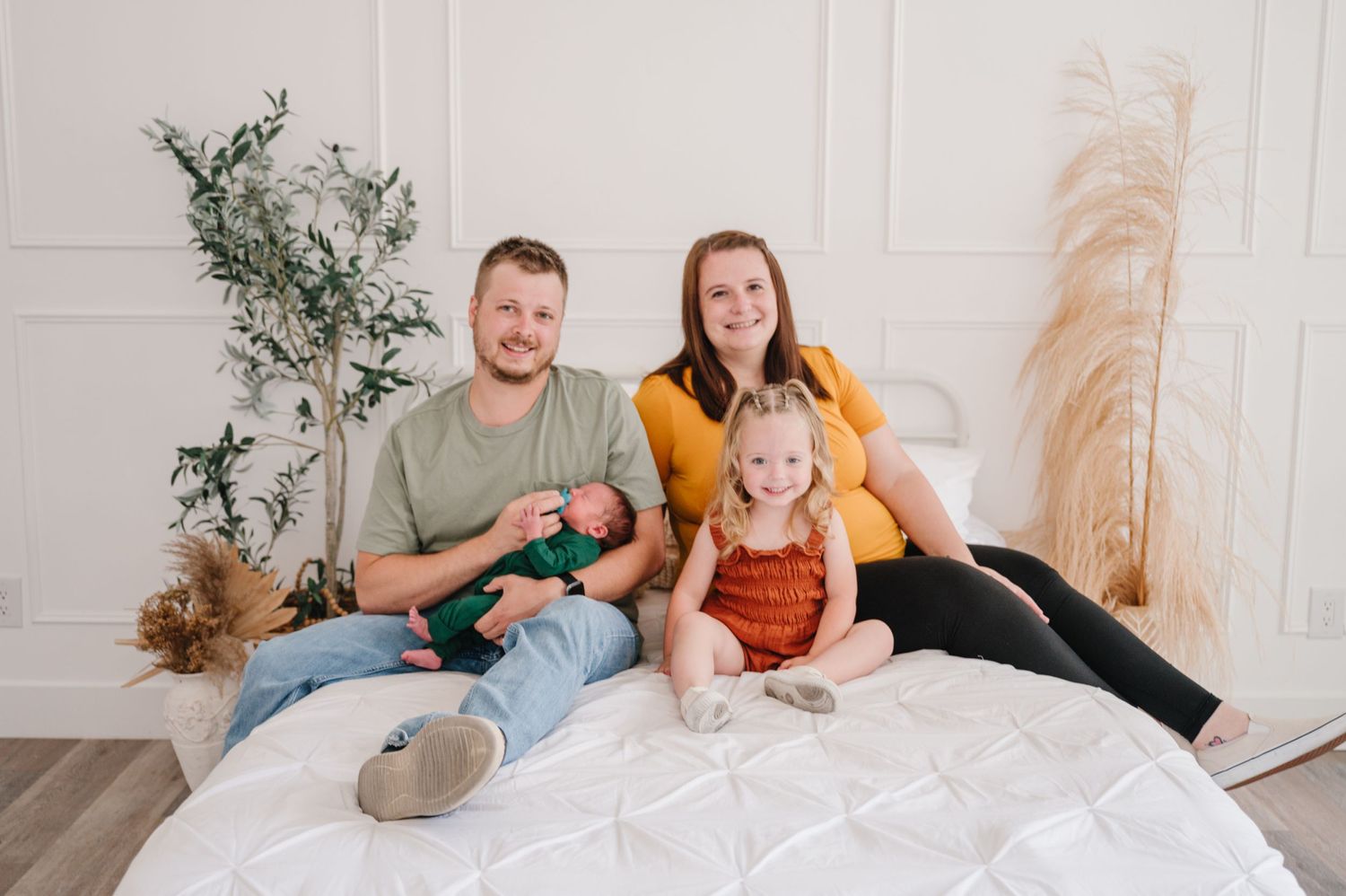 Three people sit together on a white bed with decorative plants and pampas grass in the background.