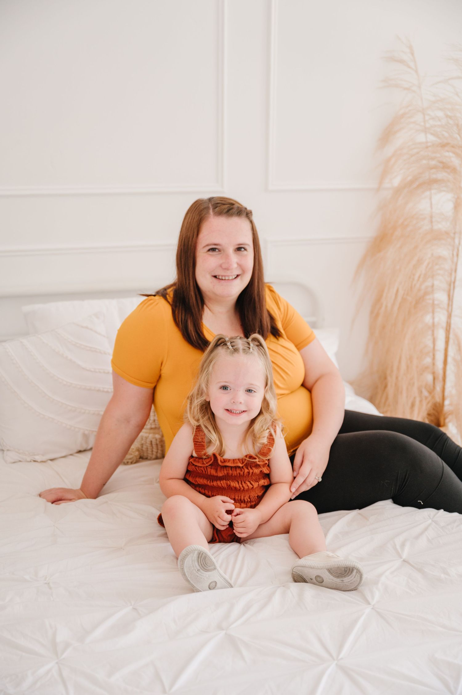 Pair sitting together on white bedding with pampas grass decor, wearing yellow and rust-colored outfits.