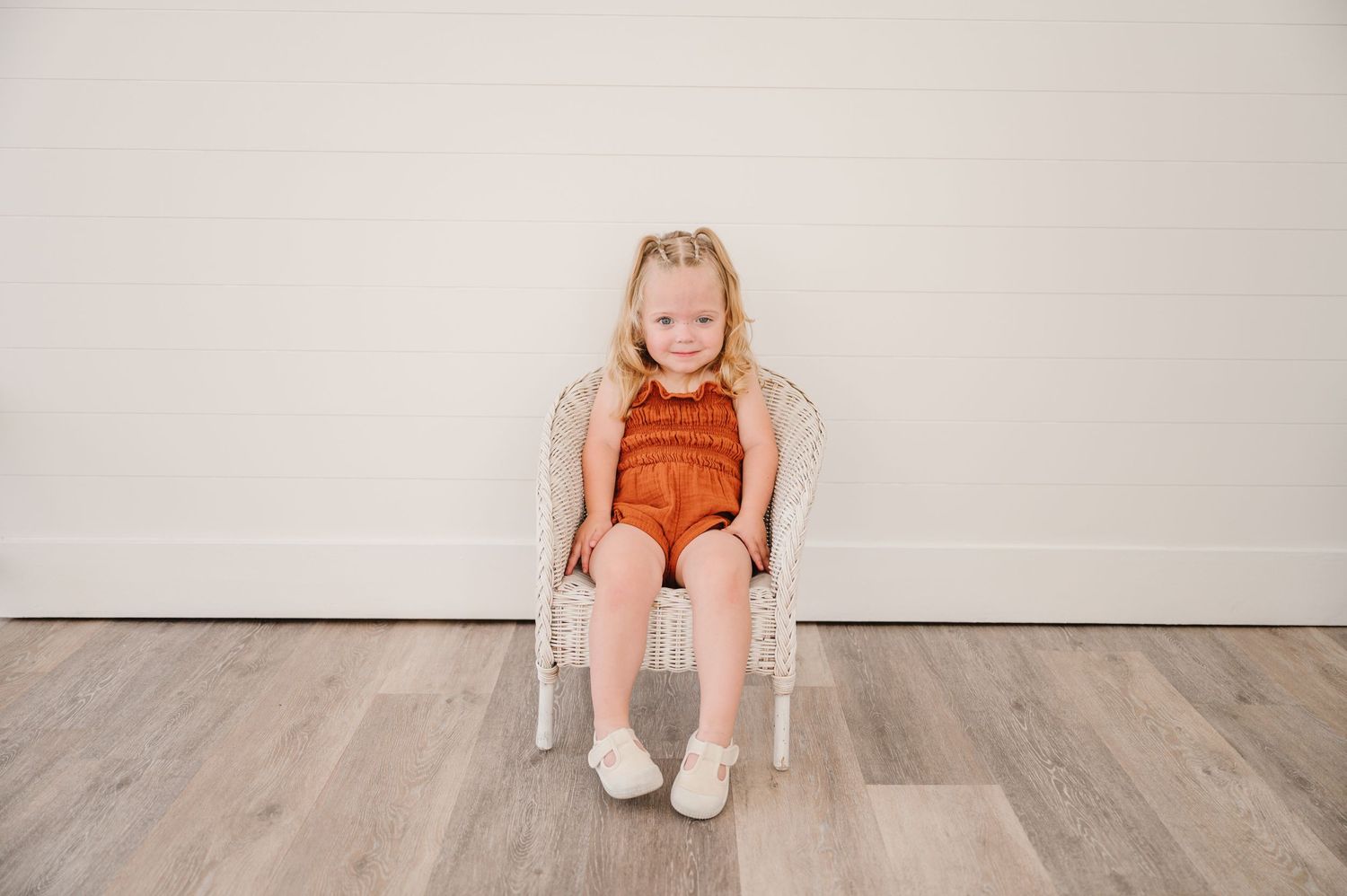 A small child in a burnt orange romper sits in a white chair against a light wall.