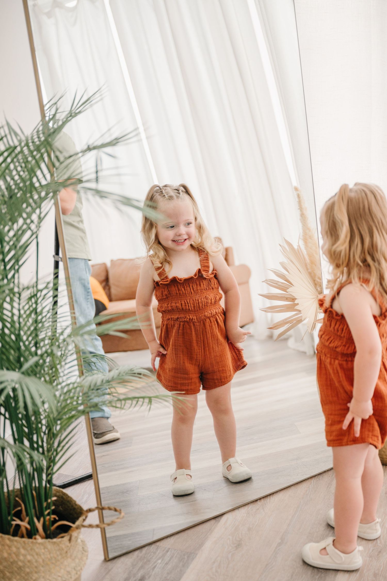 A child in an orange romper stands in front of a mirror with indoor plants and a light curtain backdrop.