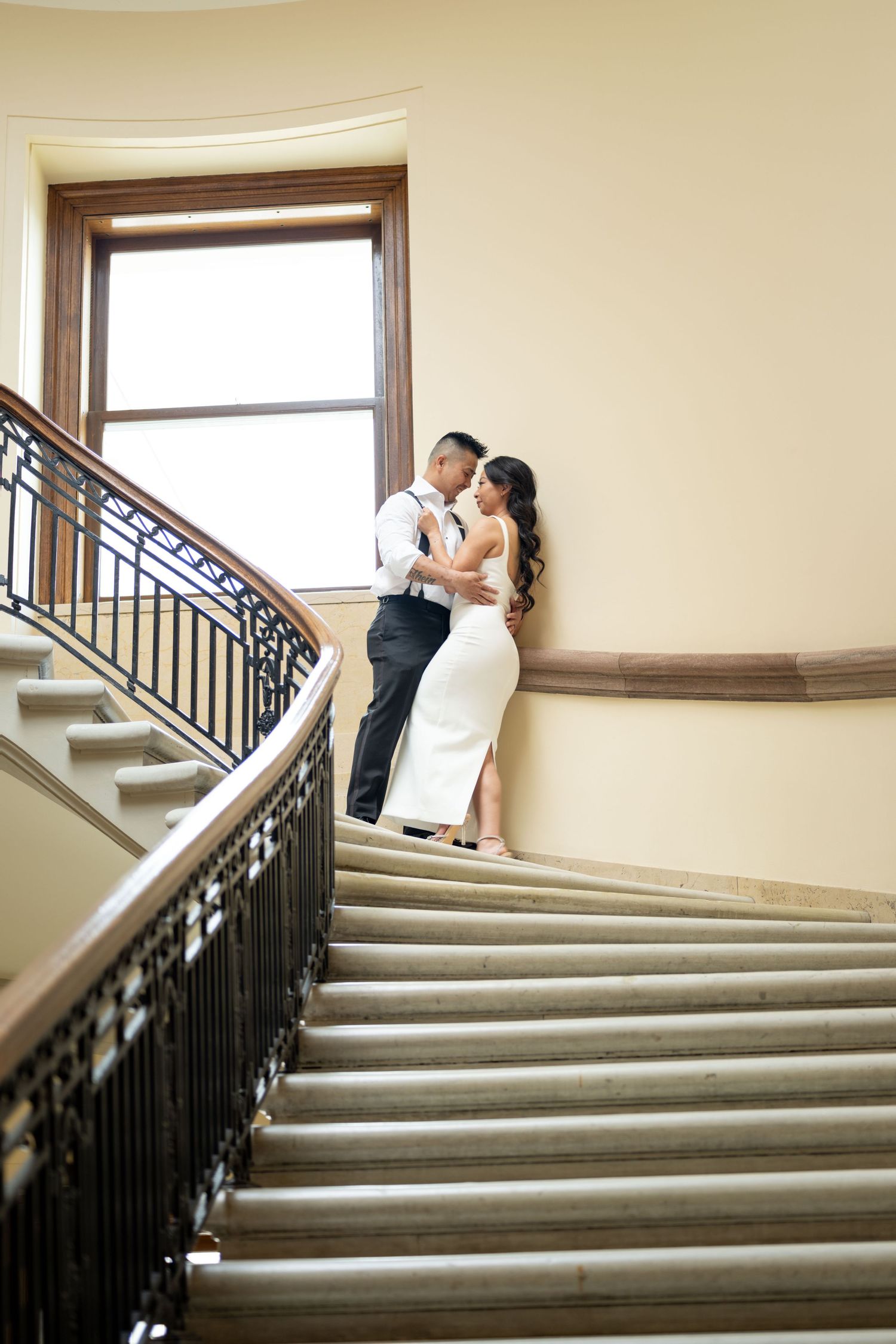 An elegant couple in formal attire sharing an intimate moment on a grand staircase with ornate railings by a window. saint Pau Photographer