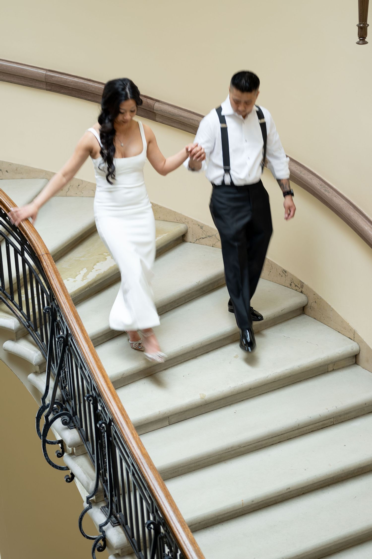Couple descends an elegant curved staircase, with bride in white gown and groom wearing suspenders. saint paul state capitol mn