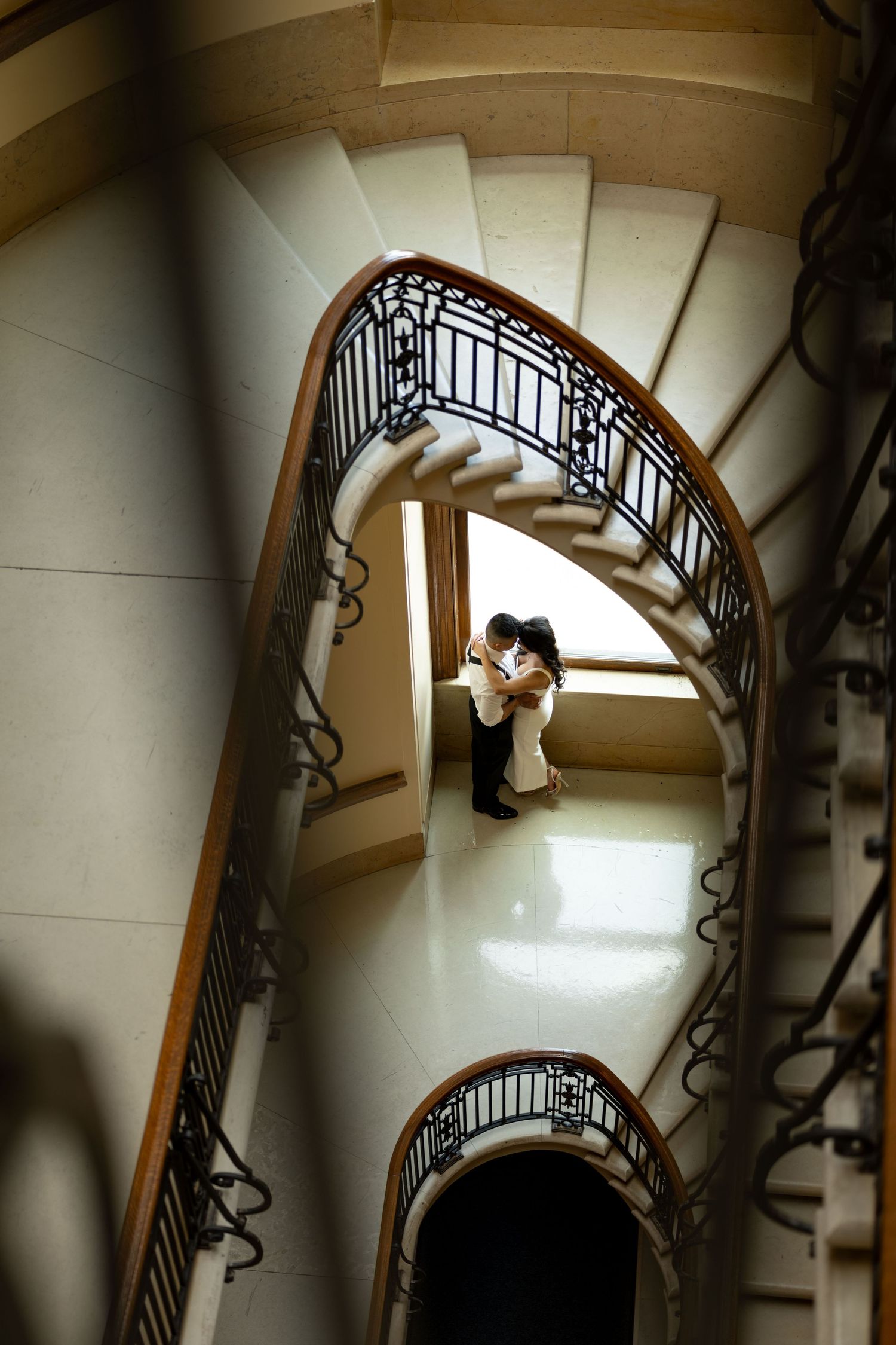 Elegant spiral staircase with ornate black railings frames a romantic couple embracing by a window in natural light. saint paul MN