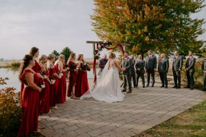 Outdoor wedding ceremony with bridal party in red dresses and groomsmen in black suits.