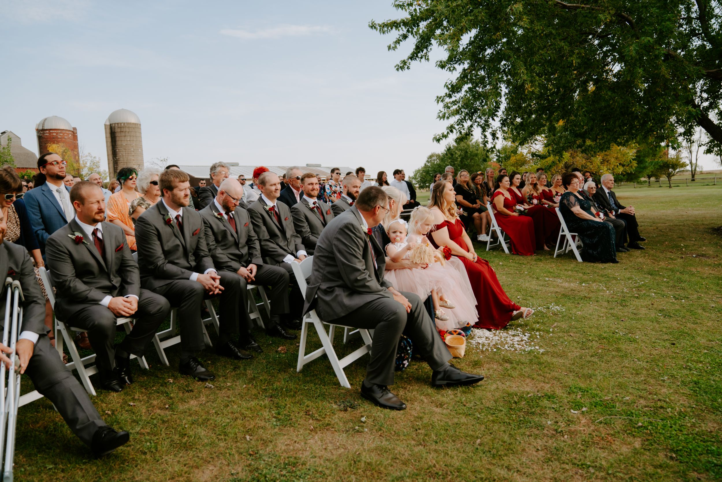 Wedding guests seated in white chairs on a grassy hillside during an outdoor ceremony.