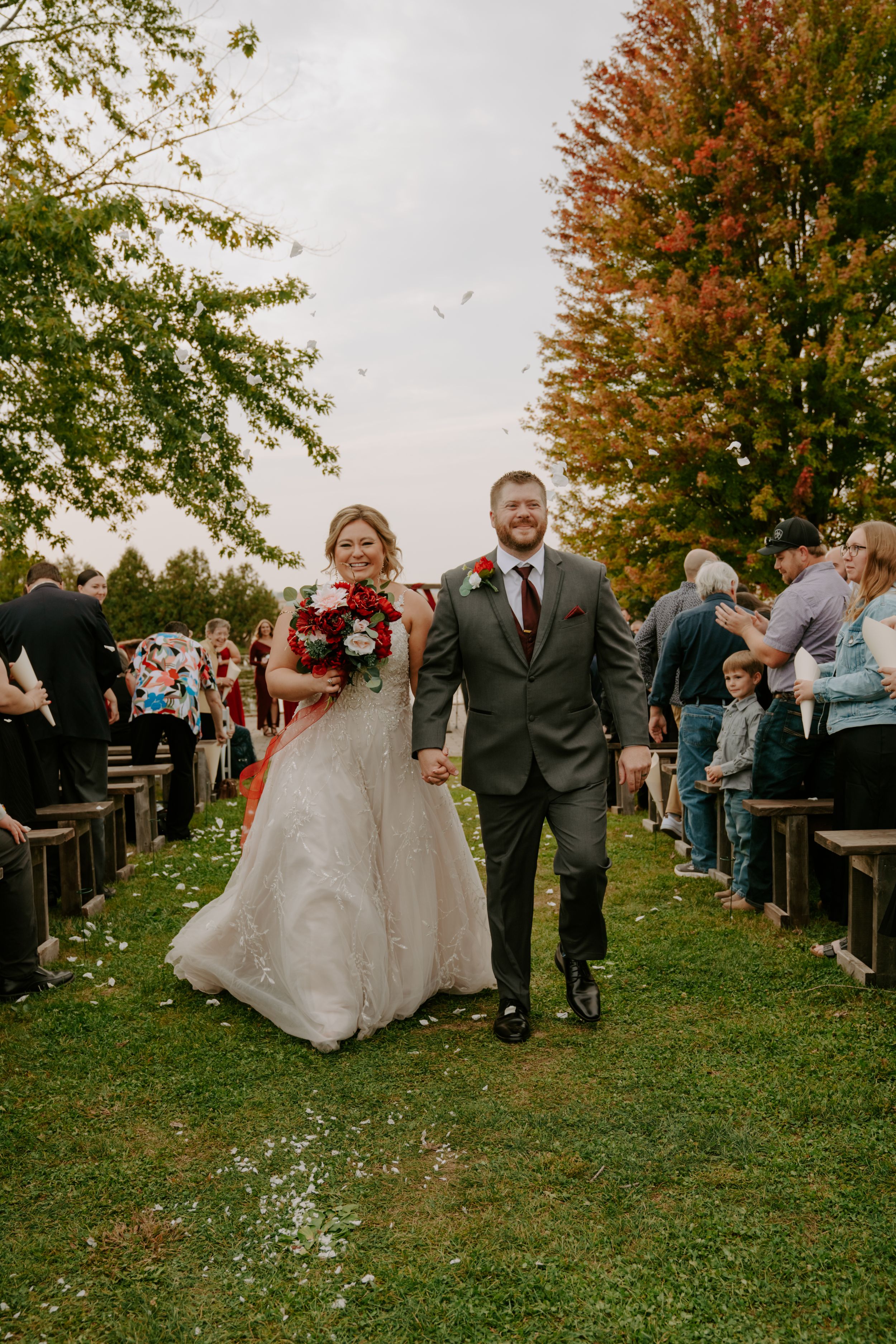 Wedding party walking down grassy aisle with autumn trees and guests seated on benches.