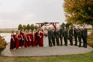 Wedding party posed outdoors with bridesmaids in burgundy dresses against fall foliage.