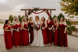 Bridal party in burgundy dresses seated together outdoors near wooden arch.