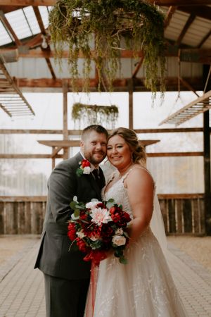 Wedding couple posing in rustic barn interior with red and white floral bouquet.