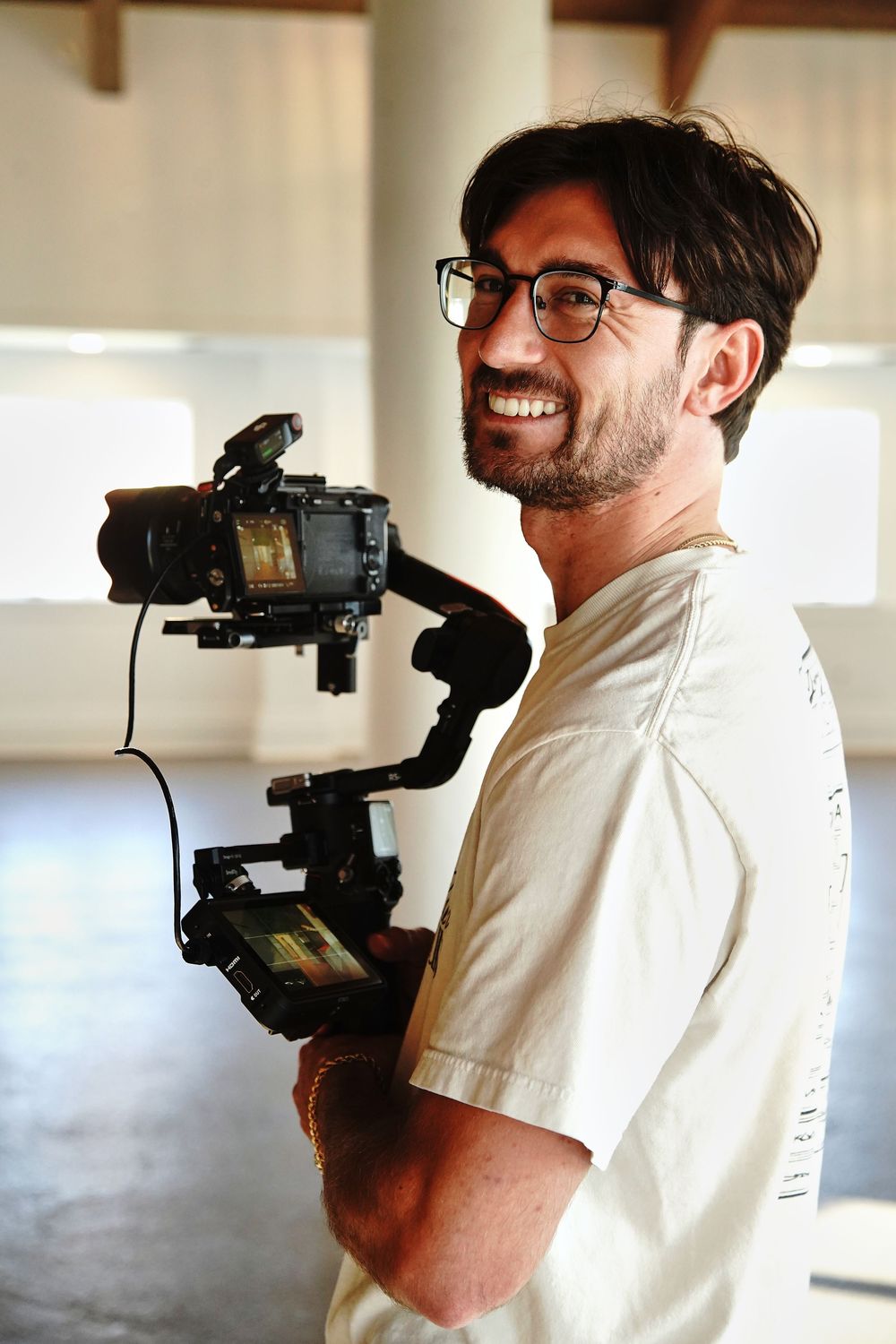 Smiling man with glasses holding a professional camera gimbal stabilizer in a bright indoor setting.