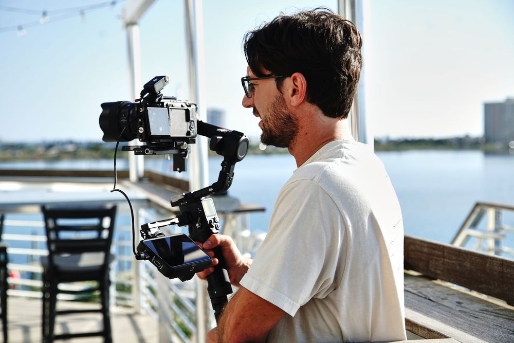 Videographer using a gimbal stabilizer to film on a sunny waterfront deck with scenic water views.