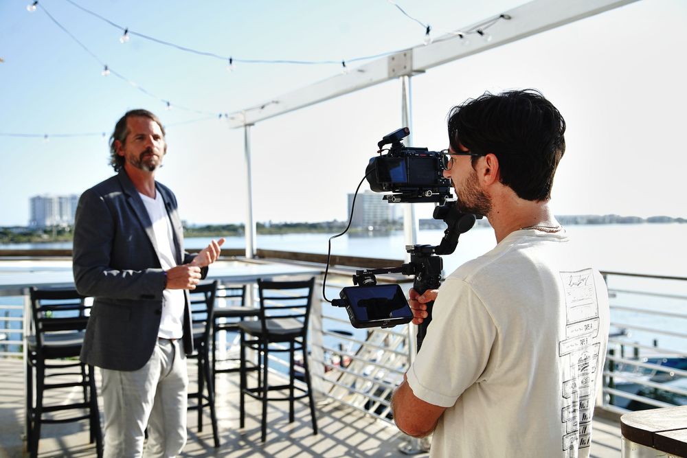 Cameraman filming a man on a waterfront deck during an outdoor video production shoot on a sunny day.