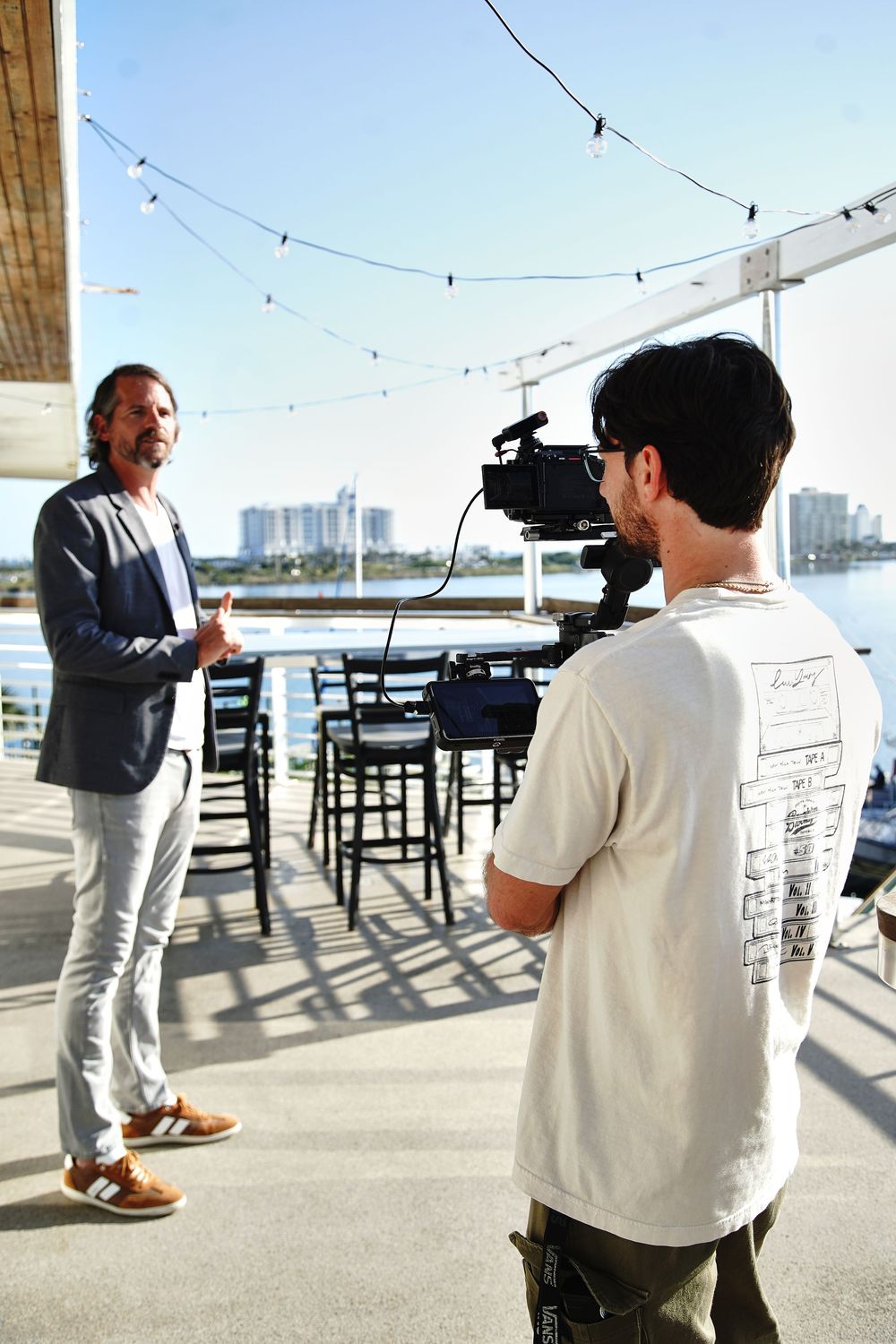 Cameraman filming a man in blazer on waterfront patio with string lights and city skyline backdrop.