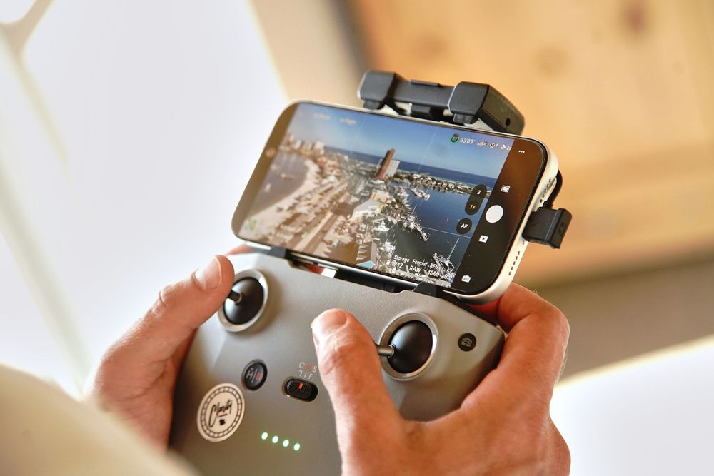 Hands holding a DJI drone remote controller with smartphone mounted showing aerial city view footage.