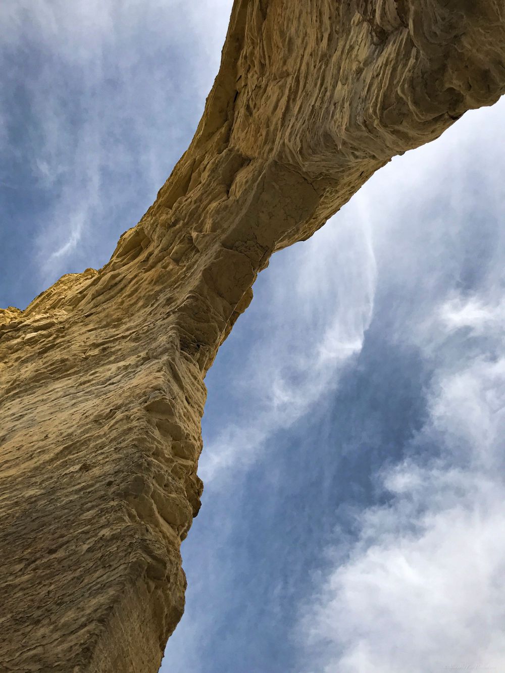 Upward view of a towering sandstone arch formation against wispy clouds in the sky.