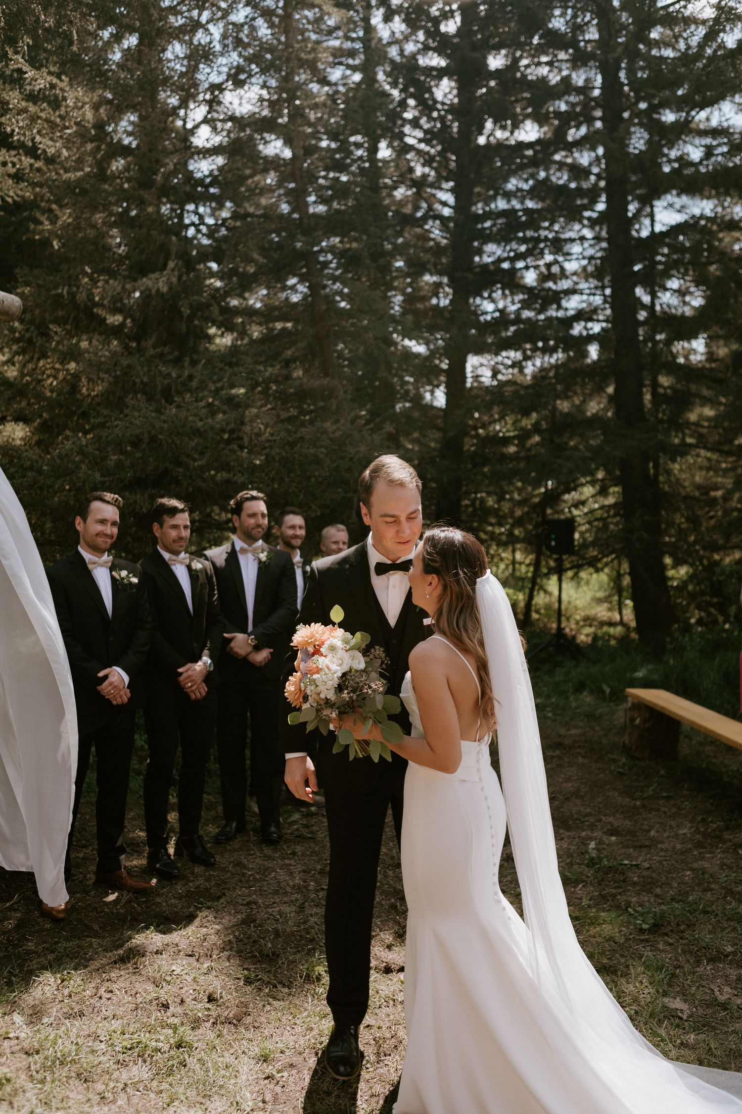 Outdoor wedding ceremony with groomsmen lined up against pine forest backdrop.