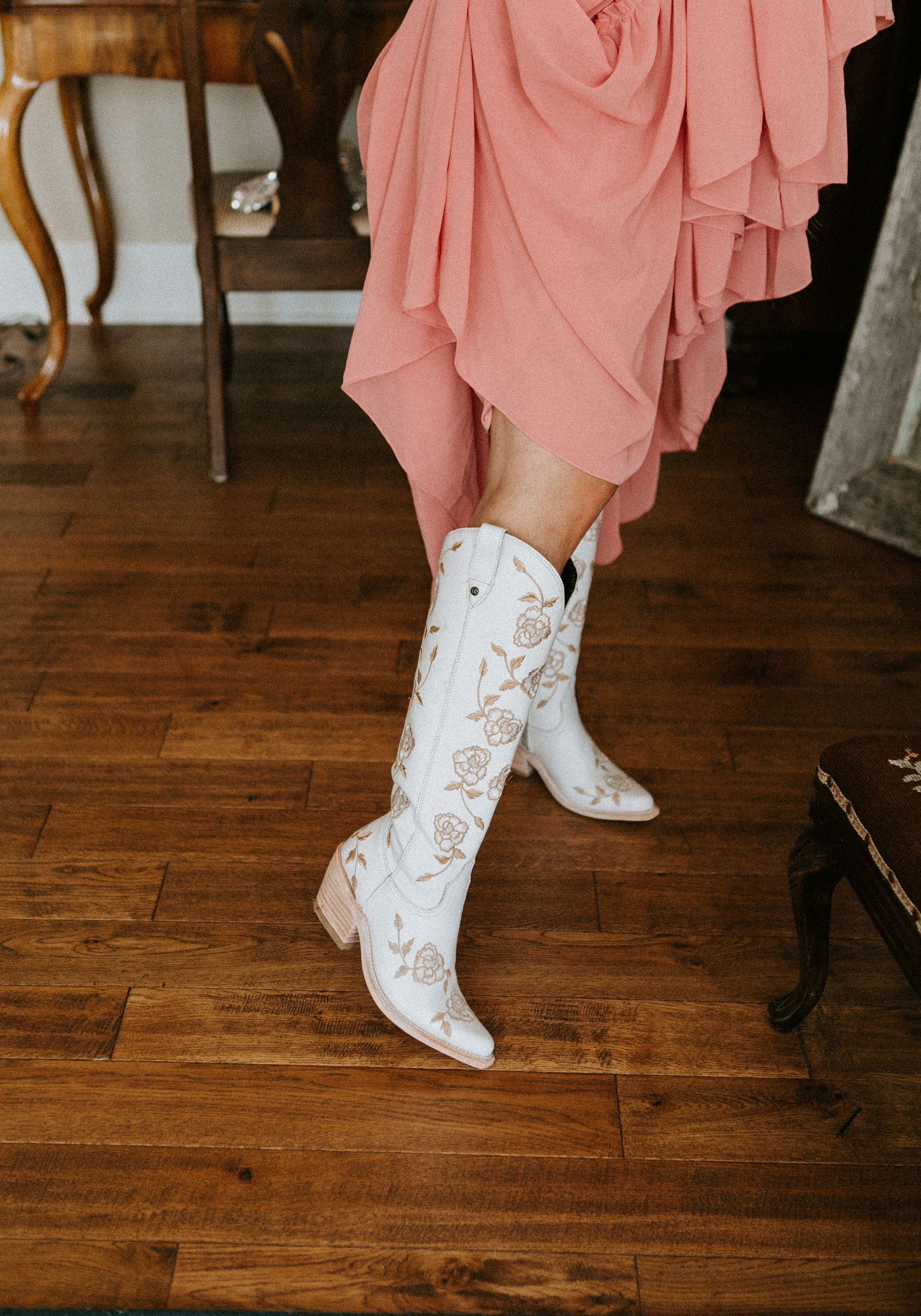 White embroidered cowboy boots with pink dress hem against wooden floor.