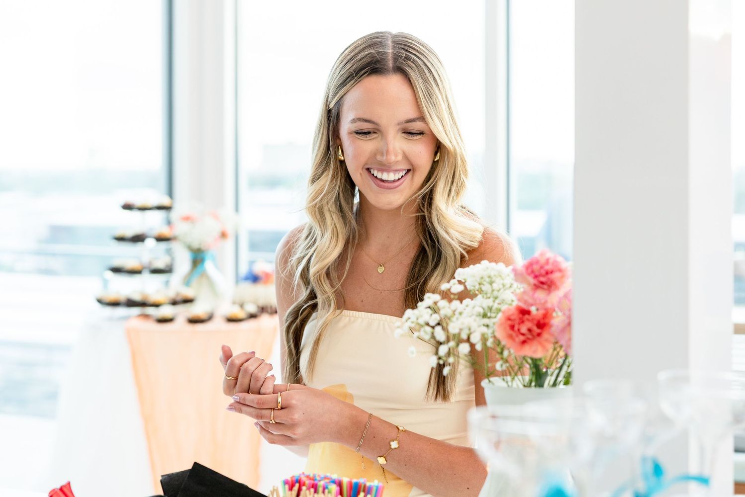 Blonde woman smiling while opening gift box at bright indoor celebration event with flowers.