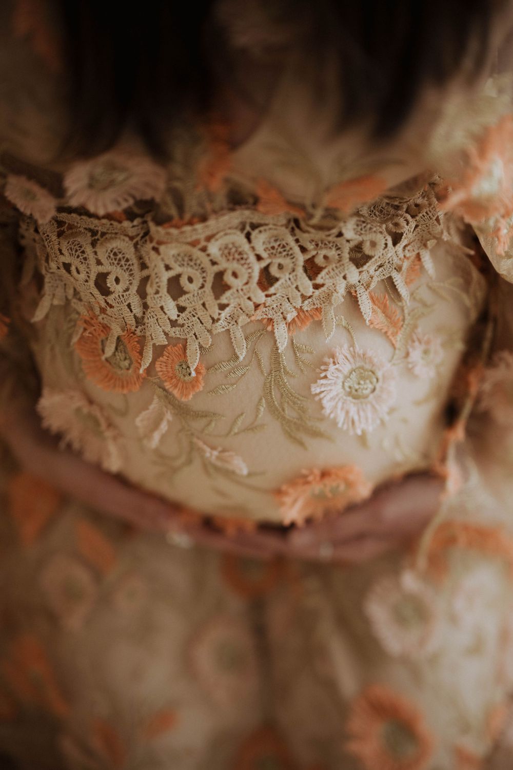 Close-up detail of vintage lace trim and delicate floral embroidery on a cream-colored fabric with soft peach tones.