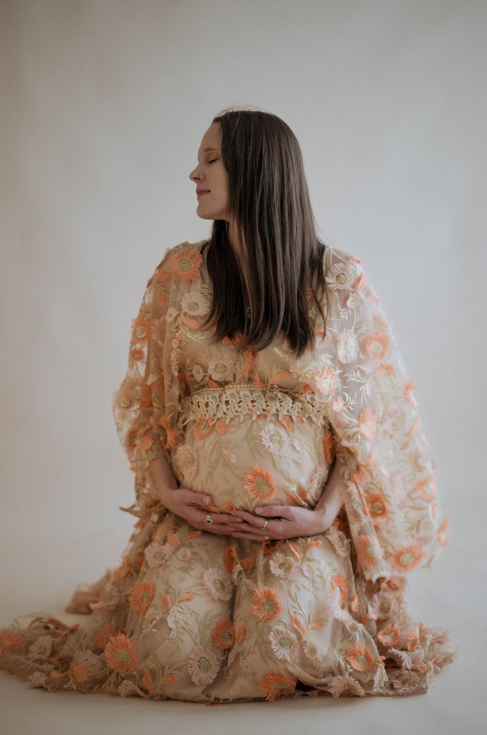 An expectant woman in a flowing peach floral dress poses elegantly in a minimalist studio setting.
