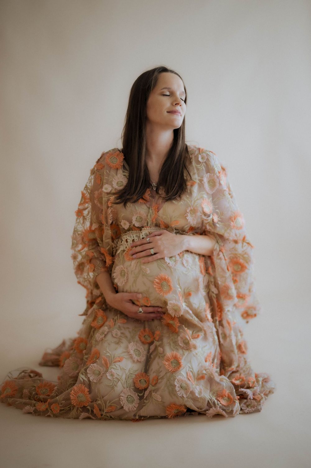 Expectant woman in floral lace dress poses serenely with hands on belly during ethereal maternity photoshoot.