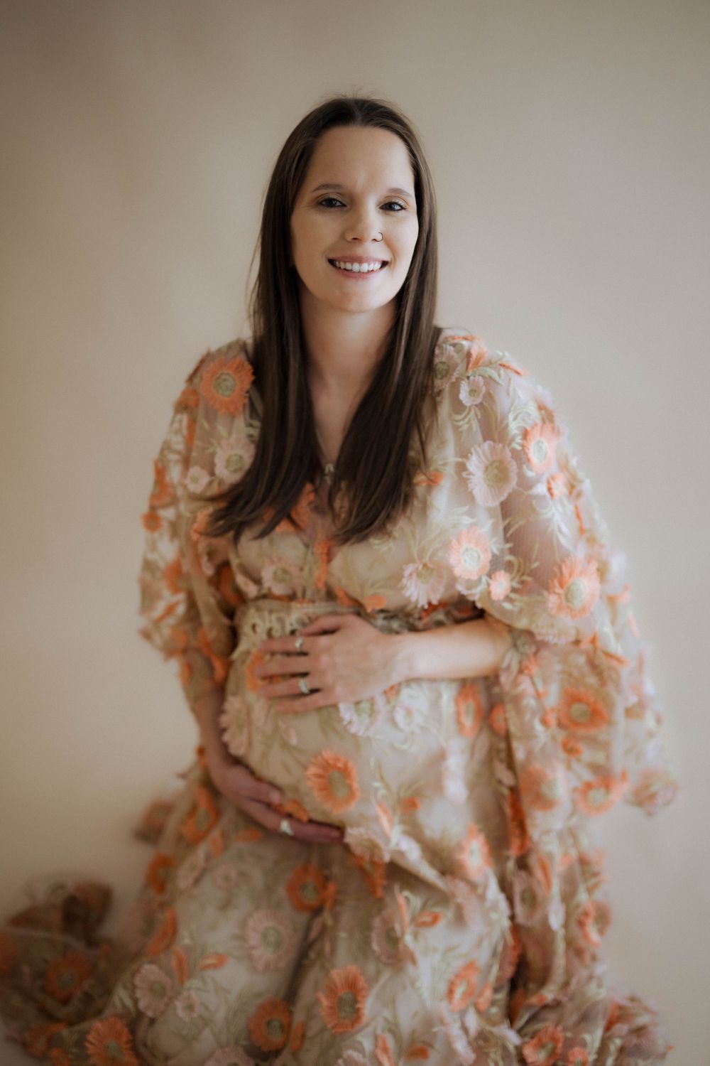 A maternity portrait featuring a woman in a floral peach dress smiling warmly while cradling her baby bump.