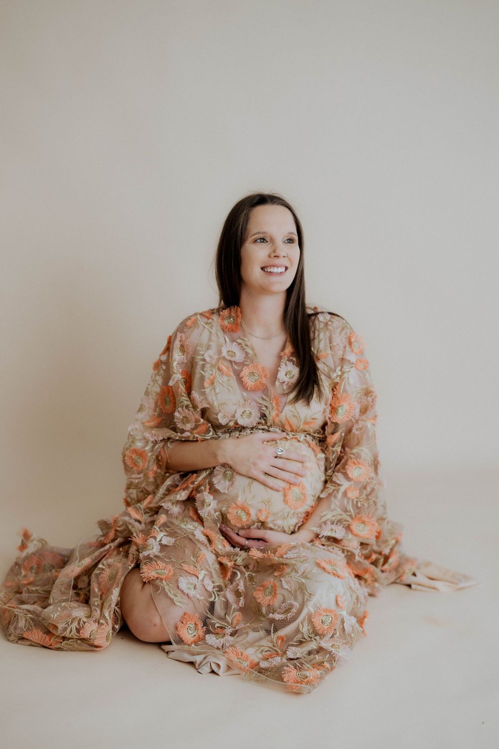 Expectant mother wearing peach floral lace dress poses on white background during maternity photoshoot.