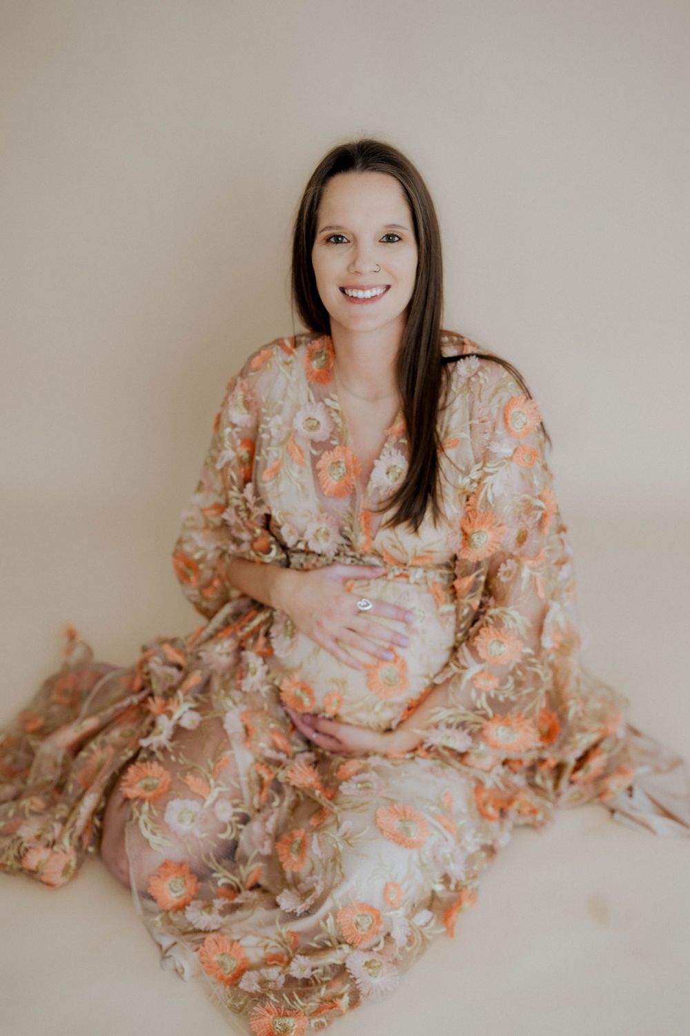 Expectant mother in floral peach and cream maternity dress sits smiling against neutral background.