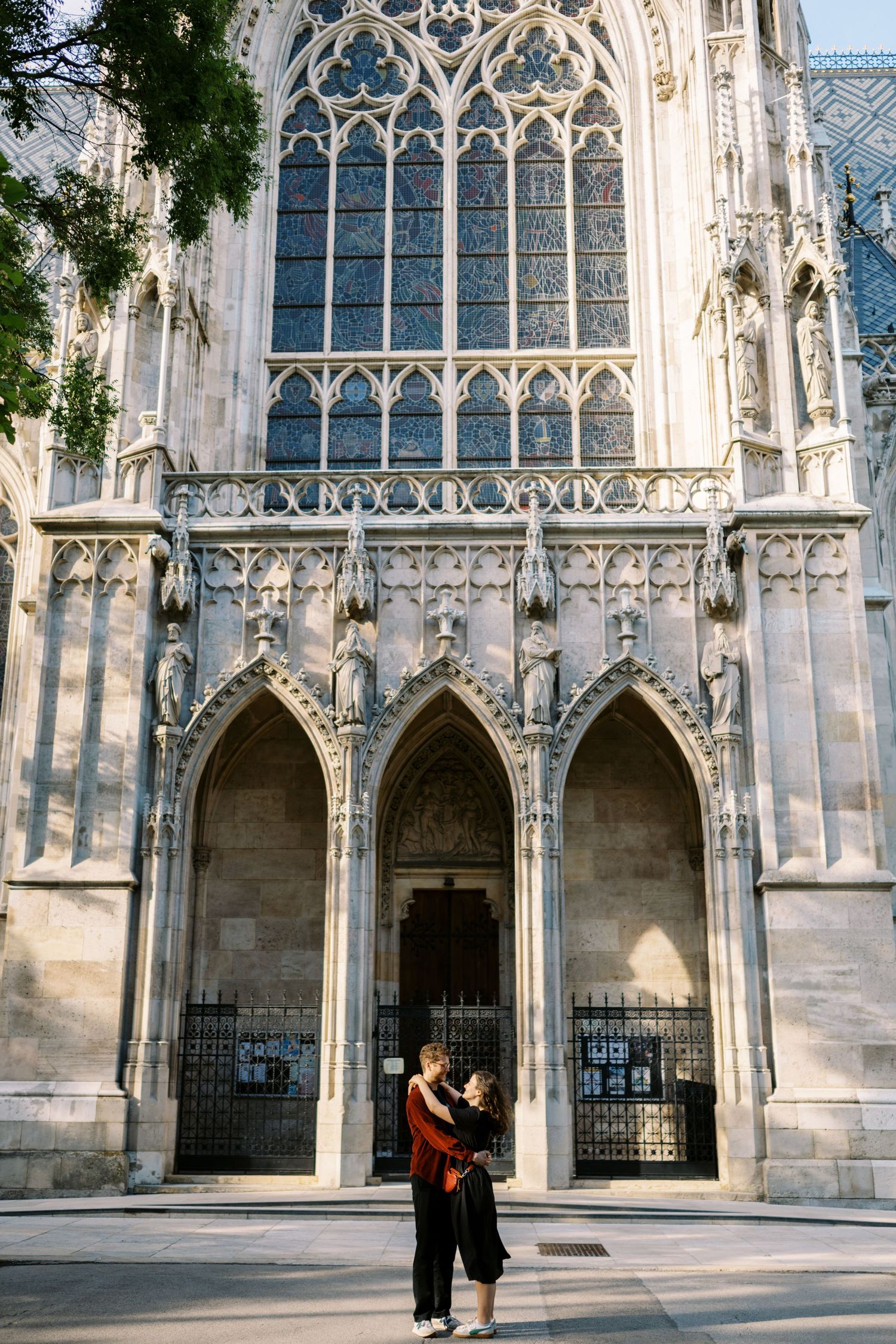 Two people embrace in front of a Gothic cathedral's ornate entrance with pointed arches and stained glass windows.