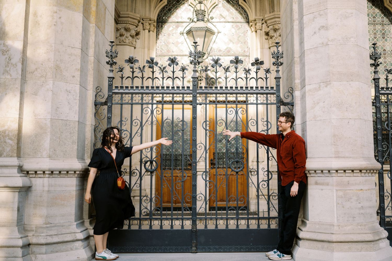 Two people pose playfully by an ornate wrought iron gate at a grand stone building entrance.