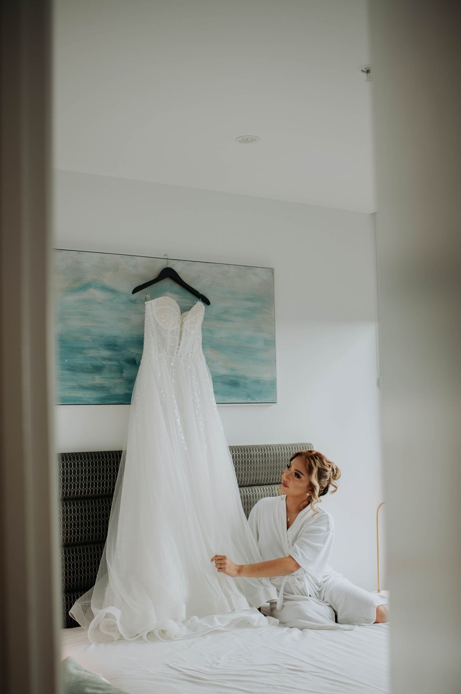 A wedding dress hangs above a bed with someone preparing nearby.