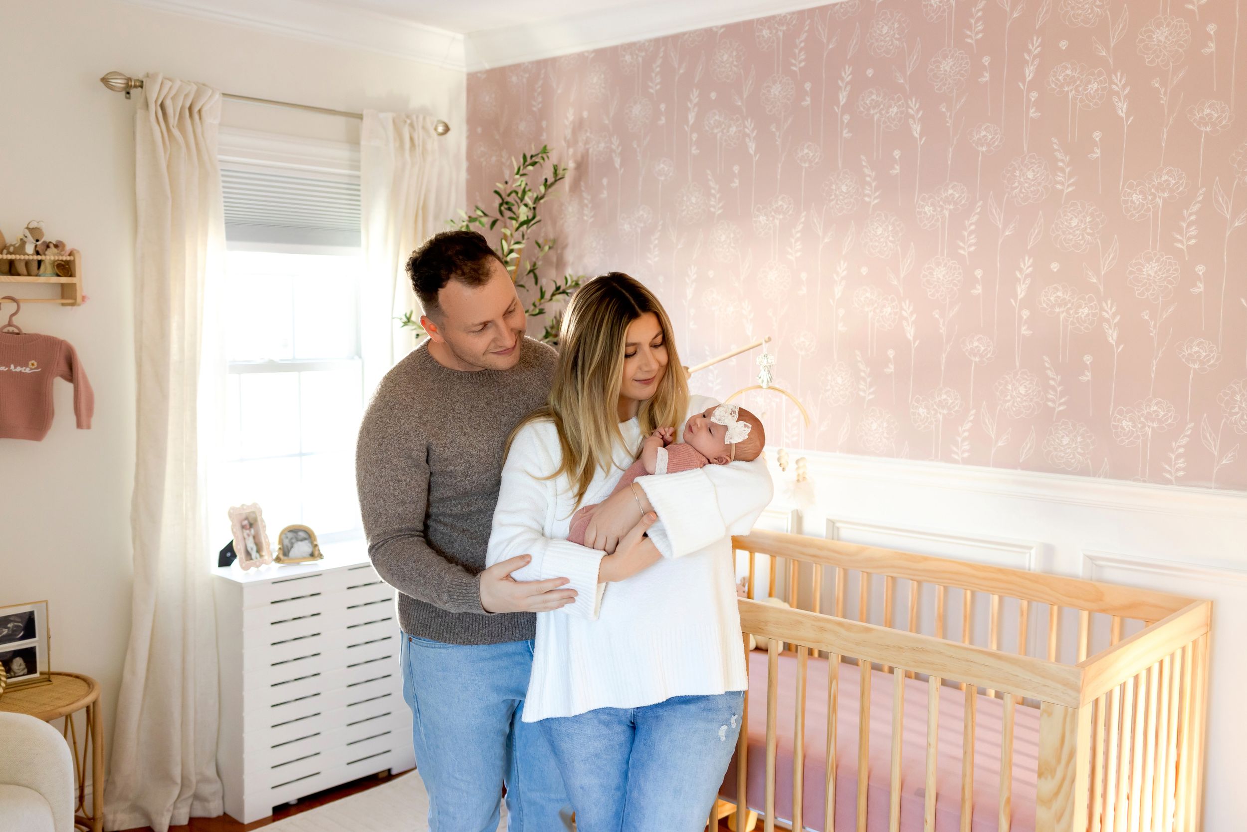 Parents standing in a pink nursery room holding their newborn baby near a wooden crib.