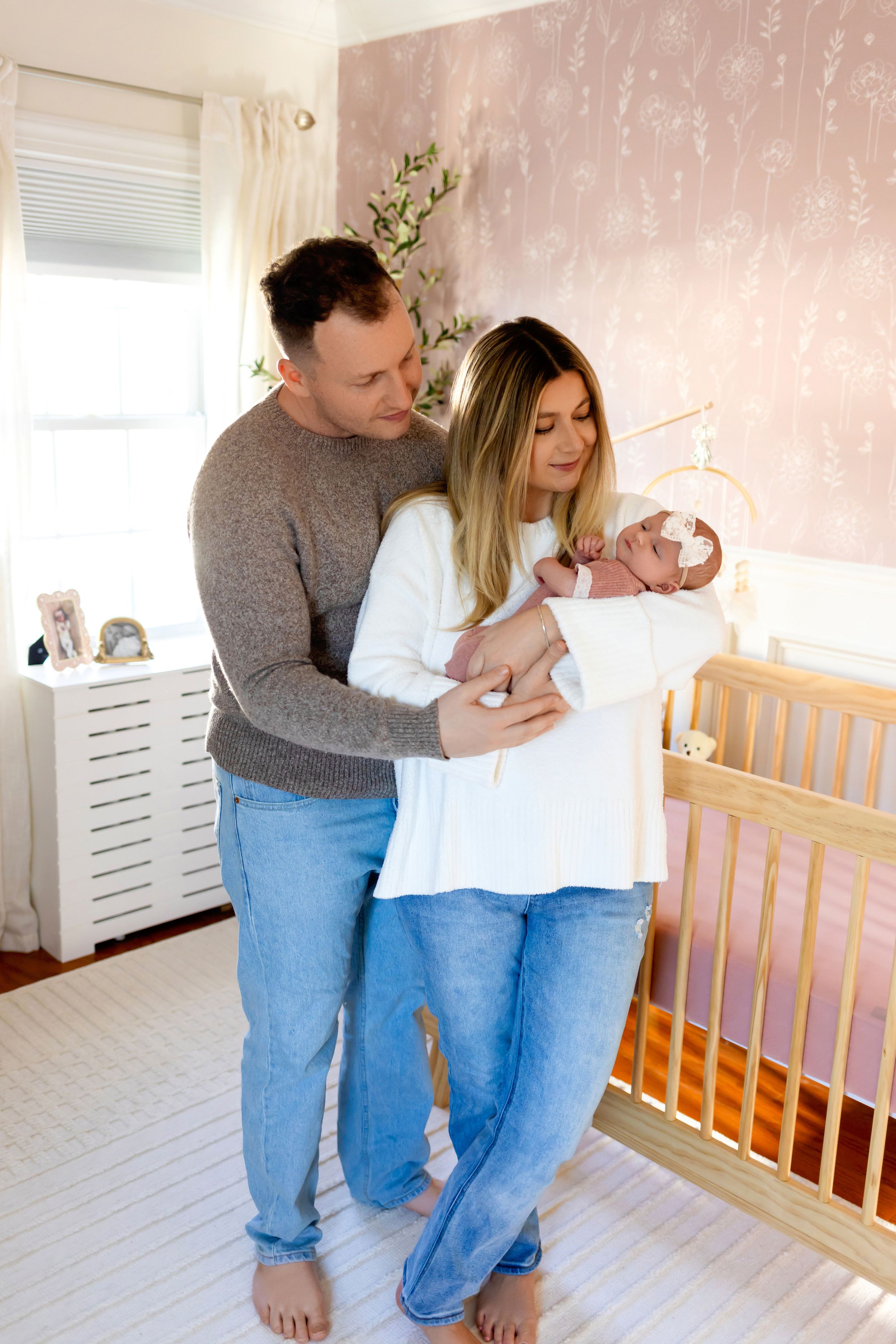 Parents hold their newborn baby in a nursery with pink floral wallpaper and wooden crib.