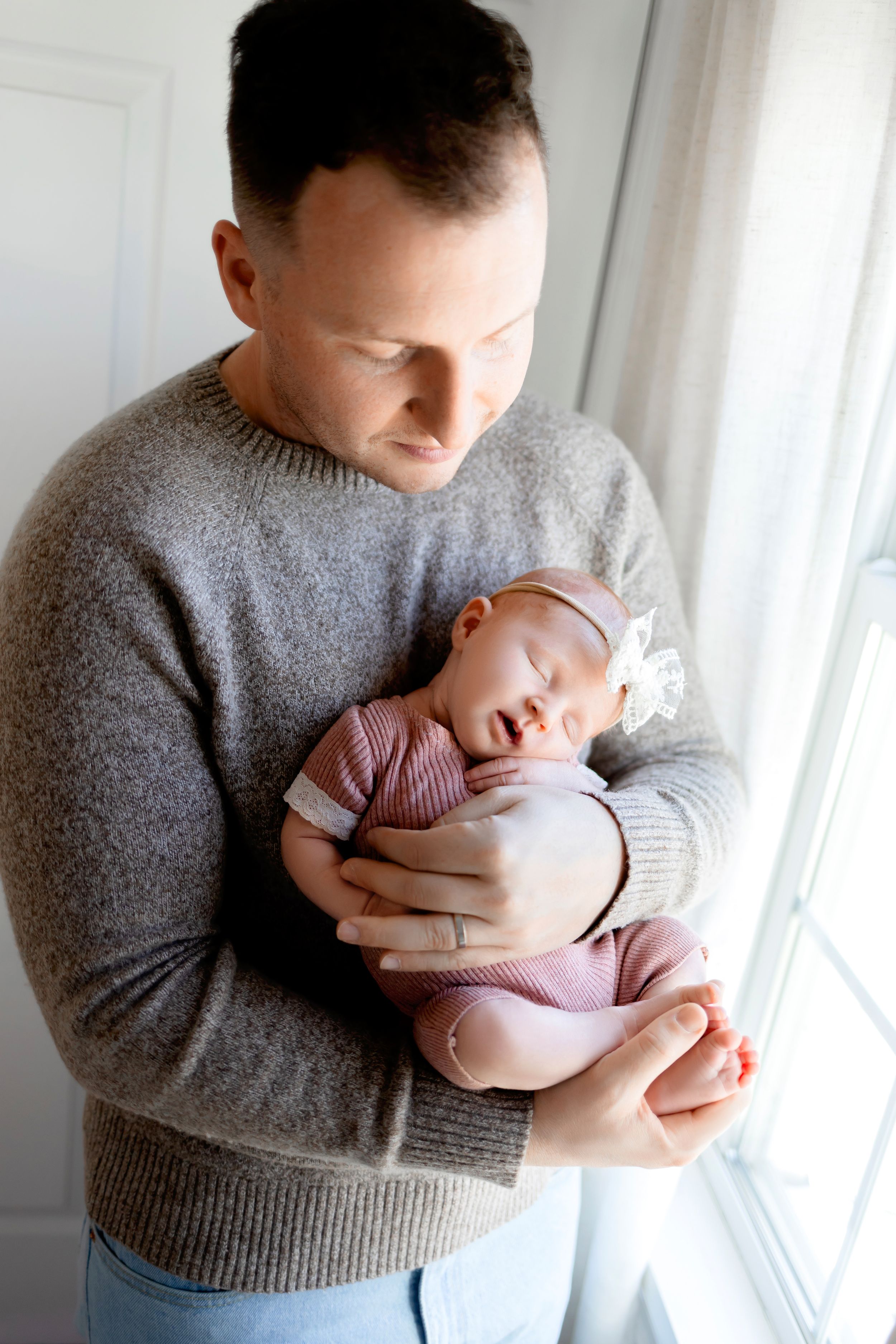 Person in grey sweater holds sleeping baby wearing pink by window.