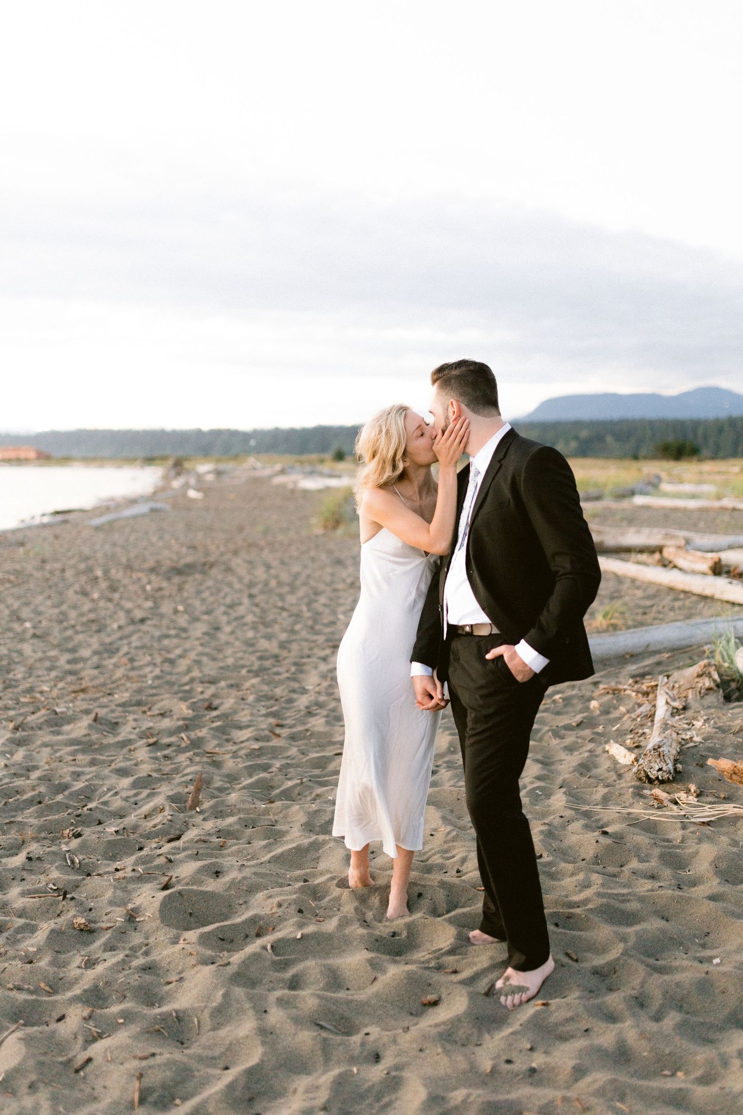 A couple shares a romantic kiss on a scenic beach with mountains in the background at sunset.