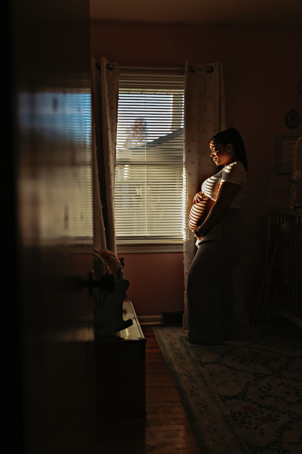 A silhouetted figure stands in a dimly lit room near a window with blinds, illuminated by soft natural light.