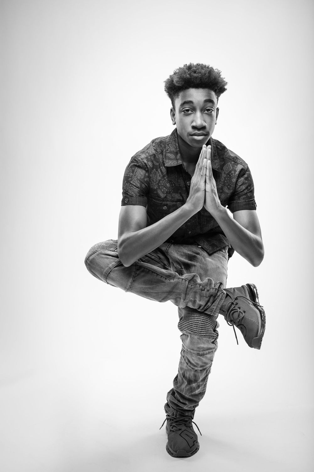Young person in casual wear strikes a balanced pose with hands pressed together in prayer position against white background.