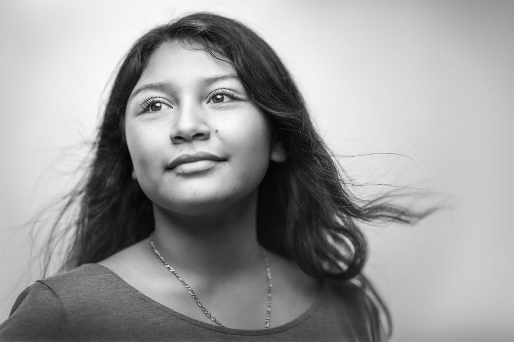 Black and white portrait of young woman with long dark hair and silver necklace gazing upward with a gentle smile.