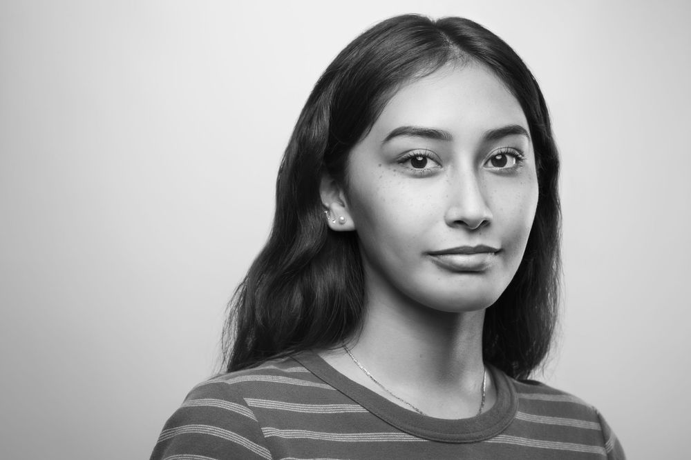 Black and white studio portrait of a young person with long dark hair wearing a striped shirt against a plain background.